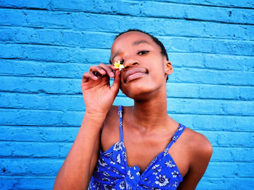 A young African girl in a floral top poses with a flower against a vibrant blue wall. Captured in South Africa.