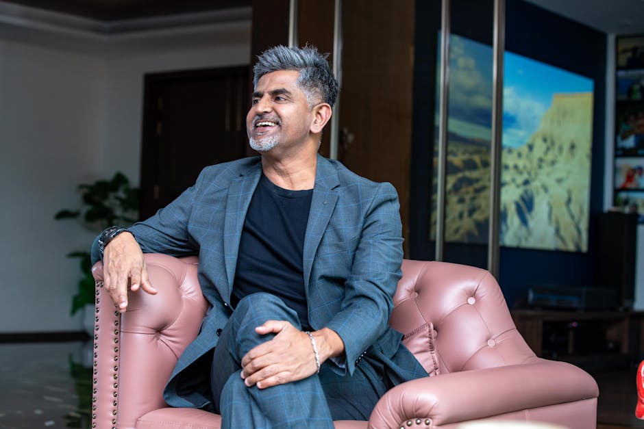 Middle-aged man in a blazer smiling and relaxing indoors on a sofa.