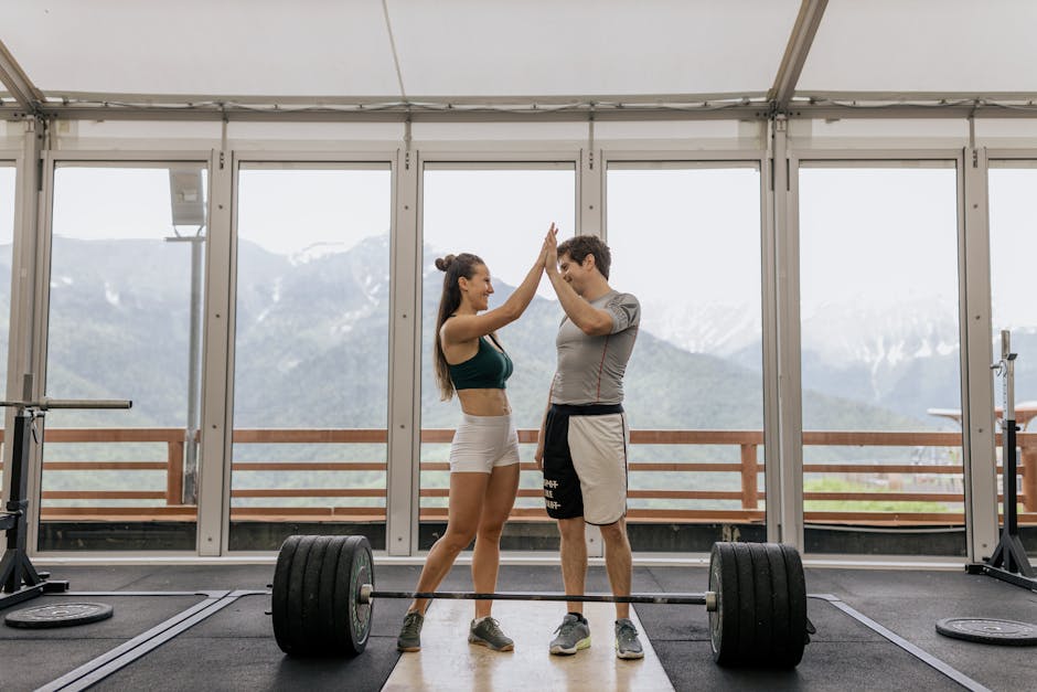 A fit couple celebrates a weightlifting success with a high-five in a bright, modern gym overlooking mountains.
