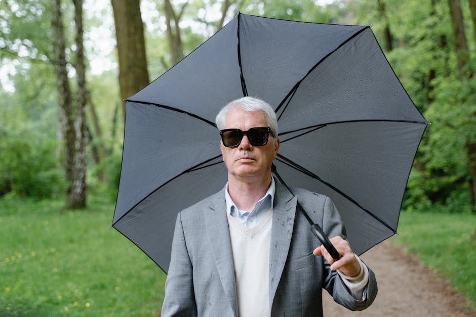 A man in sunglasses and a gray suit holds an umbrella while walking through a lush green park.