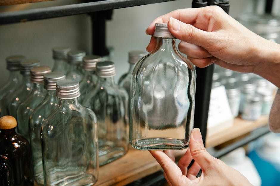 Hands holding a clear glass bottle on a shelf filled with similar bottles.