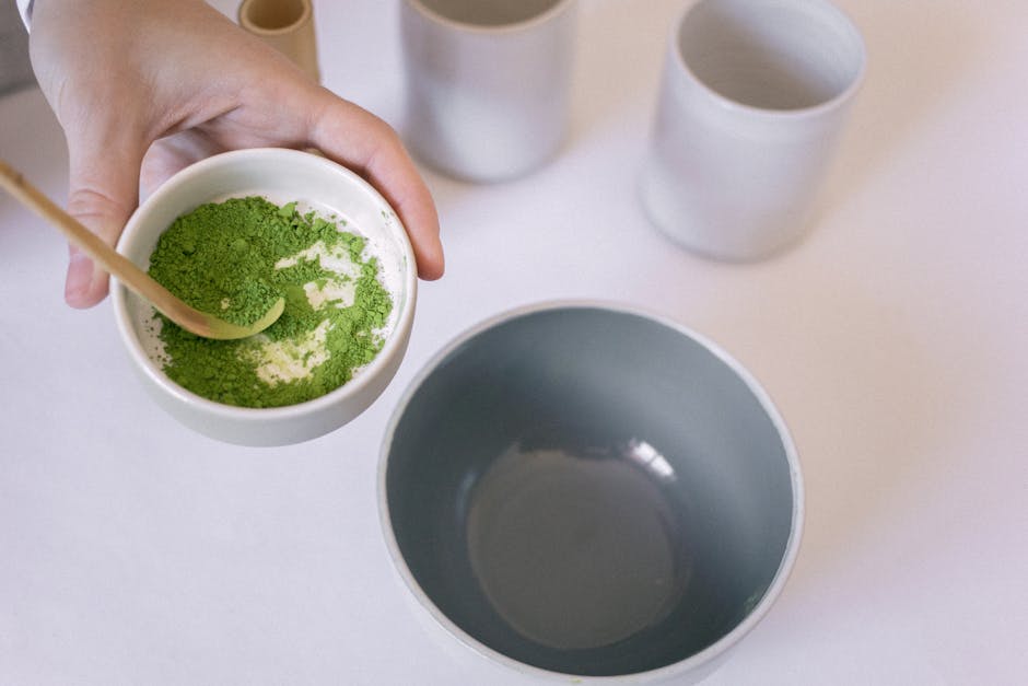 A hand holding a bowl of vibrant green matcha powder with a wooden spoon, ready for tea preparation.