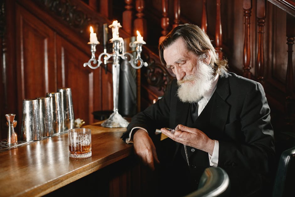 An elderly man with a beard sits at a vintage bar, holding a pocket watch with a drink beside him.