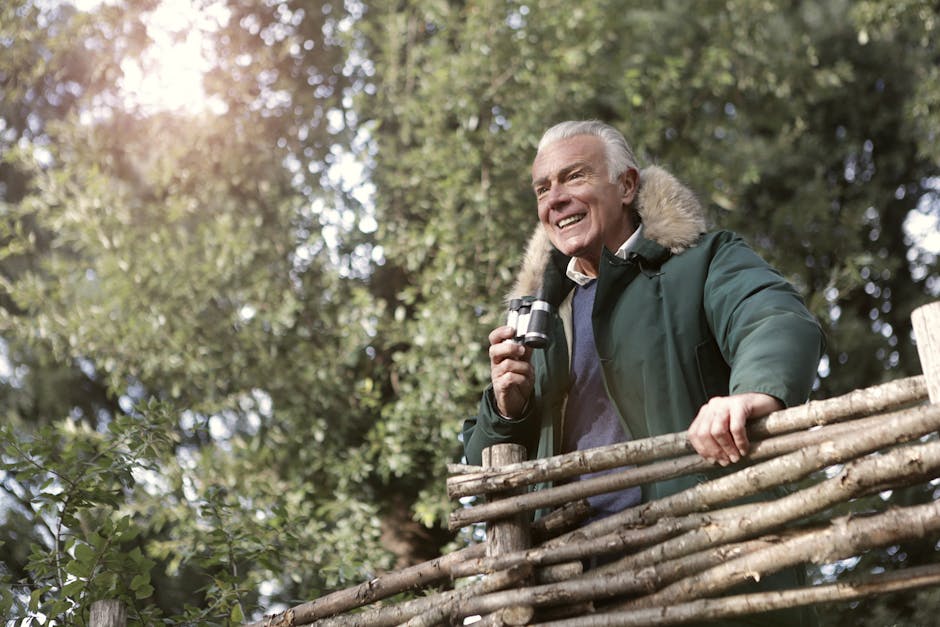 Elderly man enjoying a sunny day birdwatching in a park with binoculars.