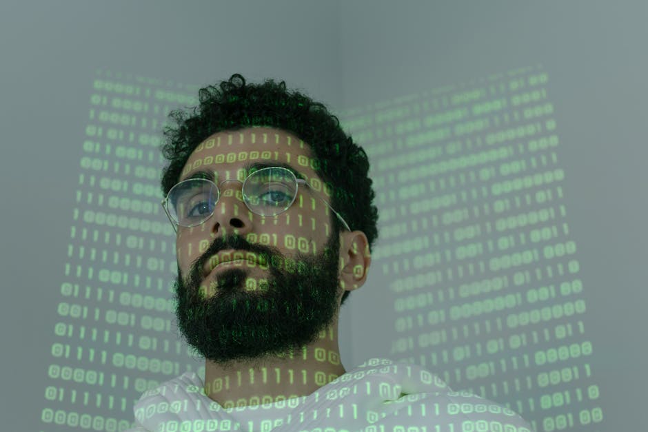 A man with a beard and eyeglasses looking at the camera, illuminated by binary code projection.