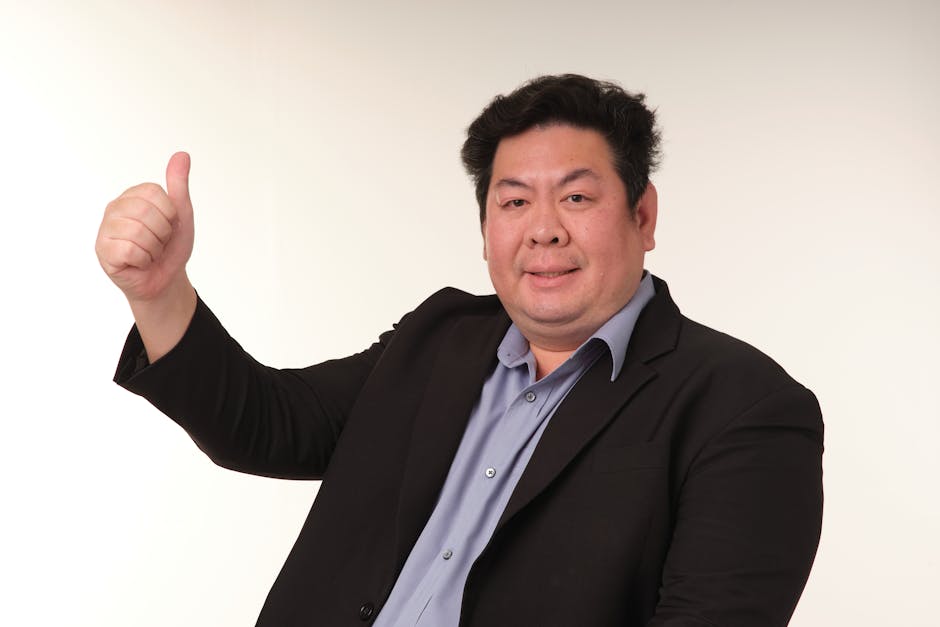 Smiling adult man in a suit giving a thumbs up gesture against a plain background.