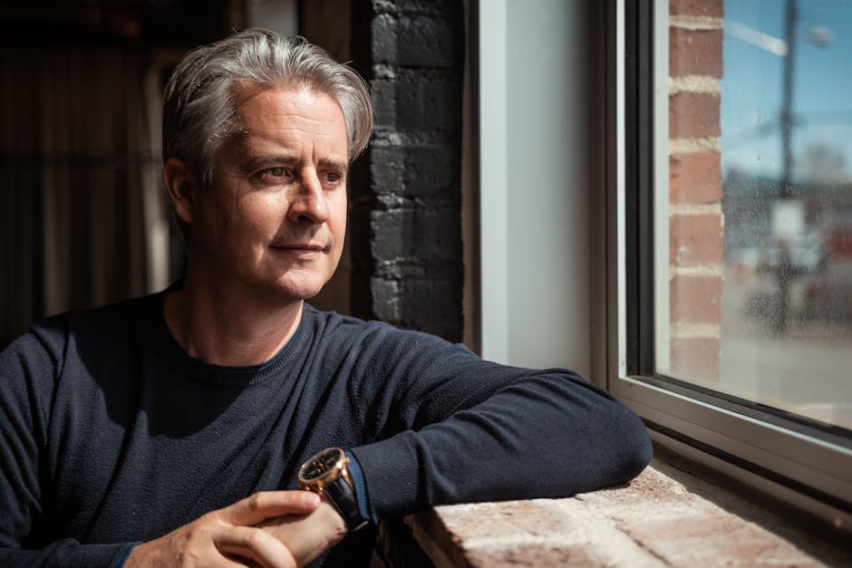 A thoughtful gray-haired man sits by a window, gazing outside during the day.